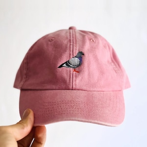 Könnte beinhalten: Eine altrosa Baseballkappe mit einer kleinen, detaillierten Stickerei einer Taube auf der Vorderseite. Der Vogel ist in Grau-, Schwarz- und Violetttönen mit leuchtend roten Füßen dargestellt, die einen Kontrast zum gedeckten Rosaton bilden.