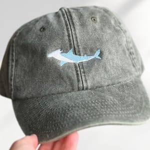 Peut inclure: Casquette de baseball grise avec un requin marteau bleu clair brodé sur le devant. La casquette a une visière incurvée et une conception classique à six panneaux, idéale pour un usage quotidien.