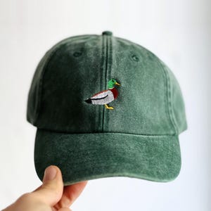 Könnte beinhalten: Eine waldgrüne Baseballkappe mit verwaschenem Vintage-Finish. Auf der Vorderseite befindet sich eine kleine, detaillierte Stickerei einer Stockente mit leuchtend grünem Kopf, weißem Halsring, brauner Brust und grauen Flügeln mit gelben Füßen.