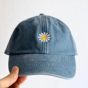 Könnte beinhalten: Eine blaue Baseballkappe mit verwaschenem Finish und einem kleinen, gestickten Gänseblümchen-Design in der vorderen Mitte. Die Blume hat weiße Blütenblätter und eine gelbe Mitte, und die Kappe verfügt über einen gebogenen Schirm mit sichtbaren Nähten.