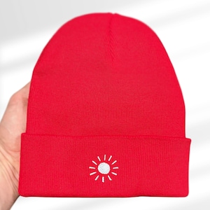 Può includere: Un berretto rosso brillante con un disegno a sole bianco ricamato sul bordo ripiegato. Il cappello è realizzato in un morbido materiale a maglia, perfetto per stare al caldo. Questo accessorio invernale è elegante.