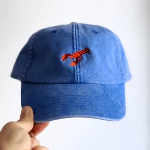 Könnte beinhalten: Eine blaue Baseballkappe mit einem kleinen, aufgestickten roten Hummer in der Mitte des Vorderteils. Die Kappe verfügt über einen gebogenen Schirm und eine Konstruktion aus sechs Paneelen mit gestickten Ösen an der Krone für eine Struktur.