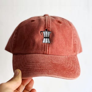 Könnte beinhalten: Eine verwaschene ziegelrote Baseballkappe mit einer kleinen, mittigen Stickerei einer silbernen Mokkakanne mit schwarzem Griff und Details. Die Kappe verfügt über eine strukturierte Krone, einen gebogenen Schirm mit sichtbaren Nähten und sechs Paneele.