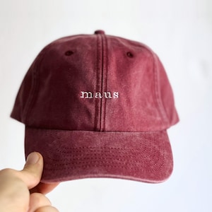 Könnte beinhalten: Eine kastanienbraune Baseballkappe mit verwaschenem Vintage-Finish. Auf der Vorderseite ist das Wort "maus" in kleinen, weißen Kleinbuchstaben eingestickt. Die Kappe hat einen gebogenen Schirm und eine Konstruktion aus sechs Paneelen mit Nähten.