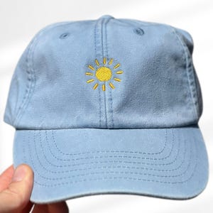 Könnte beinhalten: Hellblaue Baseballkappe mit gelbem, gesticktem Sonnenmotiv. Die Kappe hat einen gebogenen Schirm und einen verstellbaren Riemen. Ein lässiges Accessoire für sonnige Tage.