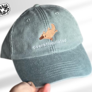 Op de afbeelding: Een saliegroene baseballcap met een bruin geborduurd vogelbekdier en het woord "overstimulated" in het wit. De cap heeft een gebogen klep en een klassiek zes-panelen ontwerp.