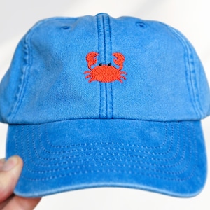 Puede incluir: Una gorra de béisbol azul con un diseño de cangrejo rojo bordado en el centro del panel frontal. La gorra tiene una visera curva, ojales cosidos para ventilación y una corona estructurada con una costura central visible verticalmente.