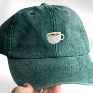 Könnte beinhalten: Eine dunkelwaldgrüne Baseballkappe mit einer kleinen, mittig platzierten Stickerei einer weißen Kaffeetasse, die mit brauner Flüssigkeit gefüllt ist. Die Kappe hat eine strukturierte Krone, einen gebogenen Schirm und sichtbare Nähte.