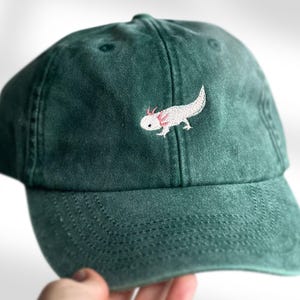 Peut inclure: Casquette de baseball vert foncé avec un axolotl blanc et rose brodé sur le devant. La casquette a une visière incurvée et une sangle réglable à l'arrière. Un accessoire décontracté et original.