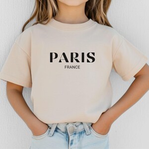 May include: A beige short-sleeved t-shirt with the word "PARIS" in black block letters and the word "FRANCE" in smaller black letters below.