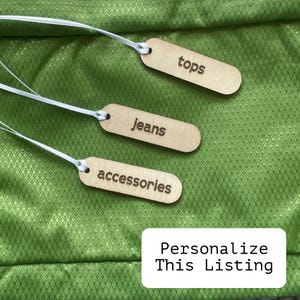 Puede incluir: Tres etiquetas de madera con una cuerda blanca atada, etiquetadas "tops", "jeans" y "accesorios". Las etiquetas están sobre un fondo de tela verde con el texto "Personalizar esta lista" en la parte inferior.