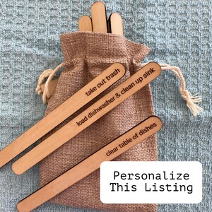 May include: Wooden sticks with the text "take out trash", "load dishwasher & clean up sink", and "clear table of dishes" written on them. The sticks are in a burlap bag.