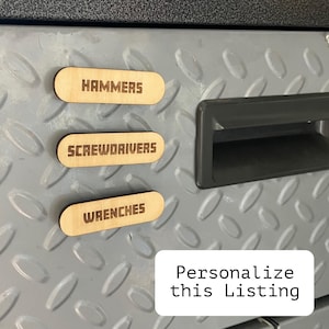 May include: Three wooden tool labels with the words "HAMMERS", "SCREWDRIVERS", and "WRENCHES" in a bold font. The labels are on a textured, gray metal surface. A white rectangle with the text "Personalize this Listing" is in the bottom right.