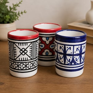 Peut inclure: Trois tasses cylindriques en céramique avec un intérieur blanc et des motifs peints à la main. Une tasse présente des lignes géométriques noires, une autre un motif floral rouge et bleu, et la troisième un motif bleu symétrique avec un bord épais.
