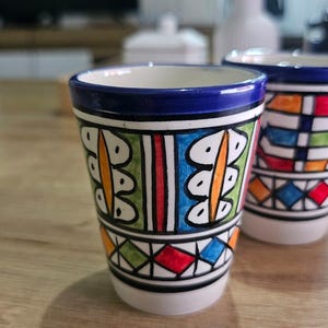 Peut inclure: Une tasse en céramique avec un motif peint à la main, un bord bleu foncé et une base blanche. L'extérieur présente des motifs géométriques en vert, orange, bleu et rouge, incluant des formes de feuilles verticales et une bande de losanges.