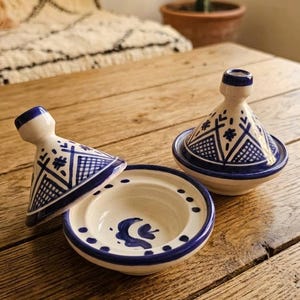 Peut inclure: Deux tajines en céramique avec un émail blanc et des motifs géométriques peints à la main en bleu. Un tajine est assemblé avec son couvercle conique, tandis que l'autre a son couvercle incliné, révélant une base circulaire avec une spirale bleue.
