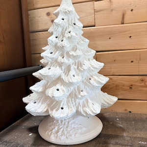 May include: A white ceramic Christmas tree figurine with a textured, branch-like surface and multiple small circular openings for lights. The tree stands on a round base decorated with embossed holly leaves and berries in a matte finish.