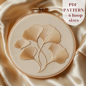 May include: An embroidery hoop with a beige fabric, showcasing a gold ginkgo leaf design. The text "PDF PATTERN + 6 hoop sizes" is in a white box. The hoop is set against a satin background.