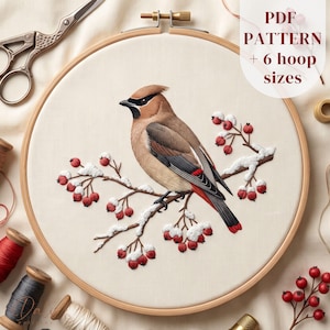 Op de afbeelding: Een borduurring met een stoffen paneel waarop een geborduurde pestvogel op een besneeuwde tak met rode bessen staat. De tekst PDF PATTERN + 6 hoop sizes is zichtbaar. Metalen scharen en diverse klossen garen liggen in de buurt.