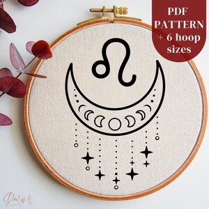 Pode incluir: Um padrão de bordado preto e branco do signo do zodíaco Leão com uma lua crescente e estrelas. O padrão é exibido em um tecido bege em um aro de bordado de madeira. O texto "PDF PATTERN + 6 hoop sizes" é exibido no canto superior direito da imagem.