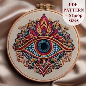 Könnte beinhalten: Gesticktes Augendesign in einem Holzreifen. Das Auge ist mit blauen, roten, gelben und goldenen Fäden detailliert. Der Text "PDF PATTERN + 6 hoop sizes" wird oben rechts angezeigt. Die Stickerei befindet sich auf einem beigen Stoff.