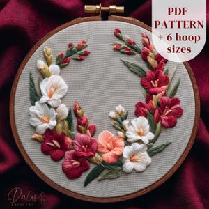Op de afbeelding: Een gedetailleerde borduurring met een bloemenontwerp met witte, rode en perzik kleurige bloemen en groene bladeren. De borduurwerk is geplaatst op een lichtbeige stof in een houten ring. Tekst op de afbeelding: "PDF PATTERN + 6 hoop sizes."