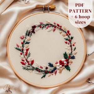May include: An embroidery hoop with a cream-colored fabric featuring a detailed floral wreath design. The wreath includes red berries, green leaves, and white and pink floral accents. The text "PDF PATTERN + 6 hoop sizes" is visible.