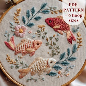 以下が含まれることがあります： 3匹のカラフルな魚と花の要素が刺繍されたフープアート。魚は赤、ピーチ、クリーム色で、青、黄色、ピンクの刺繍された葉で囲まれています。「PDF PATTERN + 6 hoop sizes」の文字が見えます。