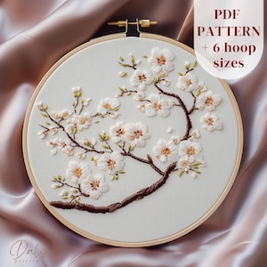 Puede incluir: Diseño floral bordado con flores blancas y ramas marrones sobre tela crema, en un bastidor de bordado de madera. El texto "PDF PATTERN + 6 hoop sizes" es visible.