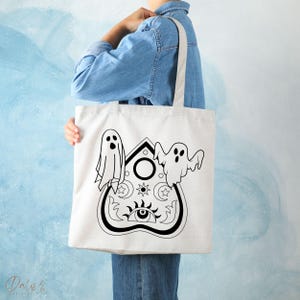 May include: A cream tote bag with a black and white Ouija board design and two ghost illustrations. The design includes stars, moons, and an eye symbol. The bag has a rectangular shape with two handles.