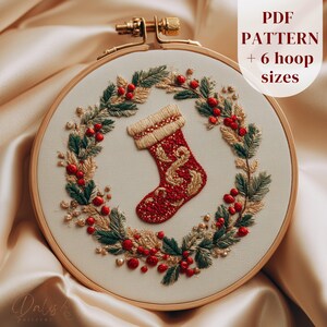 May include: Embroidered Christmas stocking in a wooden hoop. The red stocking is decorated with gold thread and surrounded by a wreath of green leaves, red berries, and gold accents. The text "PDF PATTERN + 6 hoop sizes" is in the upper right corner.