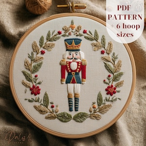 May include: Embroidered hoop art featuring a detailed nutcracker soldier in red, blue, and white, surrounded by a wreath of green and gold leaves and red flowers. The text "PDF PATTERN + 6 hoop sizes" is visible. The embroidery is set in a wooden hoop.