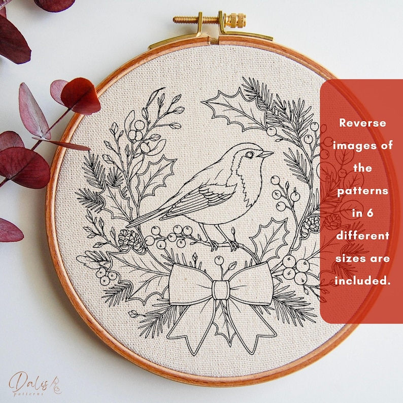 May include: A circular wooden embroidery hoop containing a cream-colored fabric with a black line art design of a bird perched within a wreath of holly leaves, berries, and pine needles, featuring a bow at the bottom and text about reverse patterns.