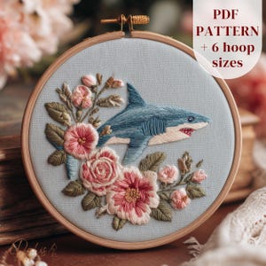 May include: Embroidered hoop art featuring a blue shark with open jaws, surrounded by pink and peach flowers and green leaves. The text "PDF PATTERN + 6 hoop sizes" is in the upper right corner. The embroidery is set in a wooden hoop.
