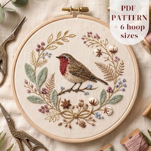 May include: An embroidery hoop containing a fabric panel with a stitched robin bird centered within a floral wreath. The design features botanical elements, berries, and leaves in muted tones. Text on the image reads: PDF PATTERN + 6 hoop sizes.