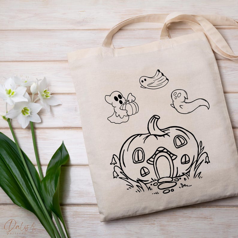 May include: Beige canvas tote bag with a black and white Halloween design. The design features a pumpkin house, ghosts, and a ghost holding a pumpkin. The bag has a handle for carrying.