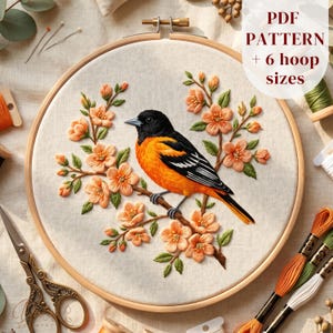 May include: An embroidery hoop containing a fabric panel with a stitched image of a black and orange bird perched on a branch with peach-colored flowers and green leaves. The text PDF PATTERN + 6 hoop sizes is visible in the upper right corner.