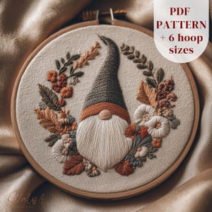 以下が含まれることがあります： 木製の刺繍枠に、ノームのデザインが施されています。ノームは灰色の帽子、白いひげ、茶色の鼻をしています。デザインには、秋色の葉と花が組み込まれています。「PDF PATTERN + 6 hoop sizes」の文字が表示されています。