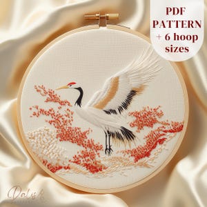 May include: Embroidered crane design in a wooden hoop. The crane has white feathers, black wing tips, and a red head. Red and orange floral accents surround the crane. The text "PDF PATTERN + 6 hoop sizes" is in the upper right corner.