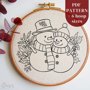 Può includere: Motivo di ricamo a linea in bianco e nero di due pupazzi di neve con agrifoglio e bacche. Il motivo è su uno sfondo di tessuto bianco in un telaio da ricamo in legno. Il testo "PDF PATTERN + 6 hoop sizes" è nell'angolo in alto a destra dell'immagine.