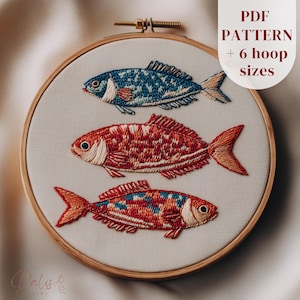 May include: An embroidery hoop featuring three colorful fish designs. The fish are embroidered in shades of blue, red, and orange, with detailed scales and fins. The text "PDF PATTERN + 6 hoop sizes" is visible. The hoop is set against a neutral background.