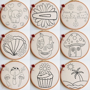 K&ouml;nnte beinhalten: Neun Stickmuster auf Stickrahmen mit verschiedenen Designs, darunter ein Pilzhaus, ein Sch&auml;del mit Pilzen, ein Cupcake mit einer Blume, eine Palme am Strand, eine Muschel mit Blumen, eine G&auml;nsebl&uuml;mchen mit einer Haarklammer, ein Elefant mit einem Herzballon und zwei Pilze.