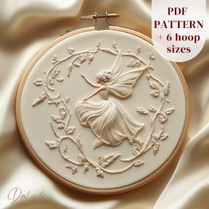 May include: Cream-coloured embroidery hoop art featuring a fairy design. The fairy has detailed wings and a flowing dress, surrounded by a floral wreath. The text "PDF PATTERN + 6 hoop sizes" is visible. The embroidery is set in a wooden hoop.