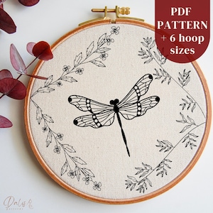 May include: A black and white embroidery pattern of a dragonfly surrounded by floral designs. The pattern is displayed in a wooden embroidery hoop. The text "PDF PATTERN + 6 hoop sizes" is displayed in the upper right corner of the image.