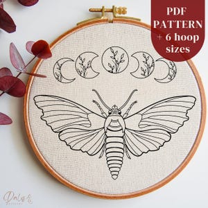Puede incluir: Un dibujo en blanco y negro de una polilla con las alas extendidas, rodeado de cuatro lunas crecientes con pequeñas ramas de hojas. El diseño está en una tela blanca estirada sobre un aro de bordado de madera. El texto "PDF PATTERN + 6 hoop sizes" está en la esquina superior derecha de la imagen.