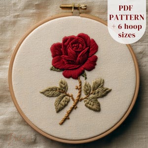 May include: An embroidered red rose with green leaves and a brown stem, displayed in a wooden embroidery hoop. The text "PDF PATTERN + 6 hoop sizes" is visible. The background is a light beige fabric, typical of craft projects.