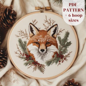 May include: Embroidered fox face in a wooden embroidery hoop. The fox is depicted with orange, white, and black fur, surrounded by green pine needles and red berries. The text "PDF PATTERN + 6 hoop sizes" is visible.