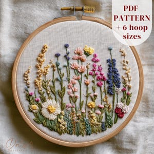 May include: A circular wooden embroidery hoop containing a fabric panel with a hand-stitched floral design featuring various wildflowers in shades of yellow, pink, blue, and white. The text PDF PATTERN + 6 hoop sizes is visible in the upper right.