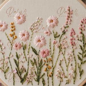 May include: Embroidered floral design featuring pink flowers with yellow centers and green stems on a cream-colored fabric. The text "Dalis Patterns" is written in a cursive font. The embroidery is set in a round wooden hoop.