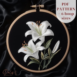Puede incluir: Un bastidor de bordado con un diseño detallado de lirios blancos sobre tela negra. El bastidor de madera tiene un cierre de latón. El texto "PDF PATTERN + 6 hoop sizes" es visible. El diseño incluye hojas y tallos verdes.